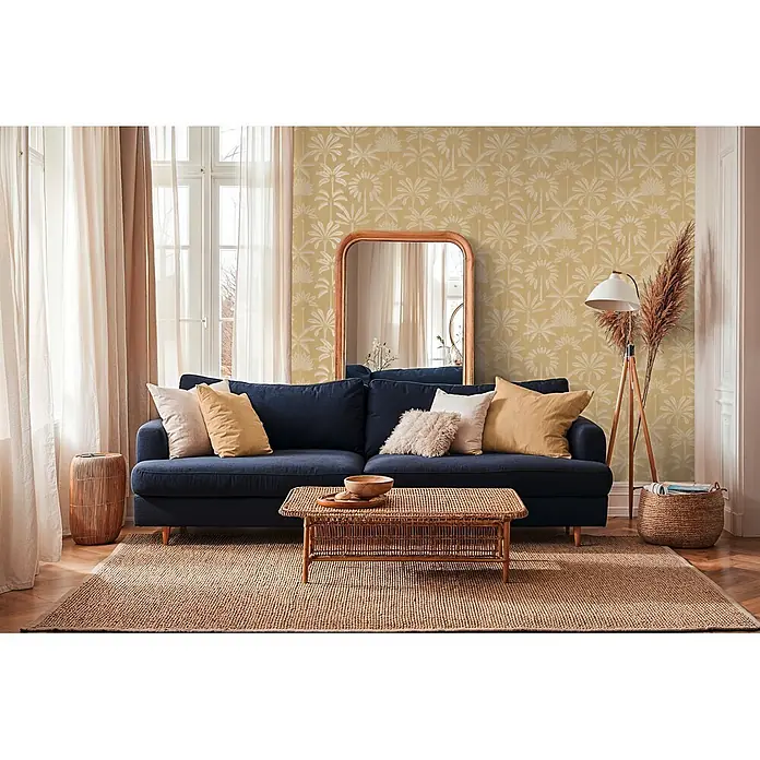 A.S. Création Cuba 2 Vliestapete Palmen II - Braun-Creme, Floral, 10,05 x 0,53 m - Ein marineblaues Sofa mit neutralen Kissen, ein geflochtener Couchtisch und ein Jute-Teppich in einem gemütlichen Wohnzimmer mit gemusterter gelber Tapete.