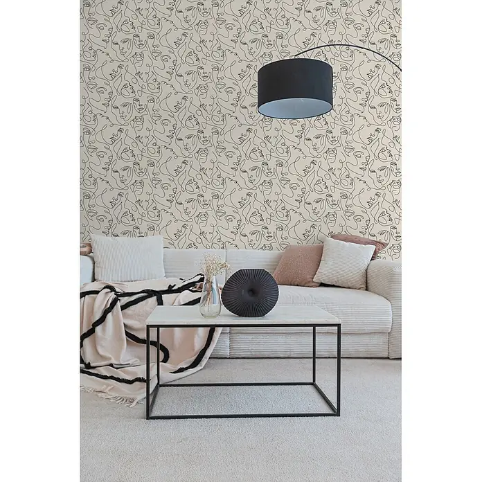 A.S. Création Pintwalls 3 Vliestapete Gesichter - Weiß-Schwarz, Grafisch, 10,05 x 0,53 m - Beige Tapete mit schwarzem abstraktem Gesichtermuster dekoriert eine Wohnzimmerwand hinter einem Sofa.