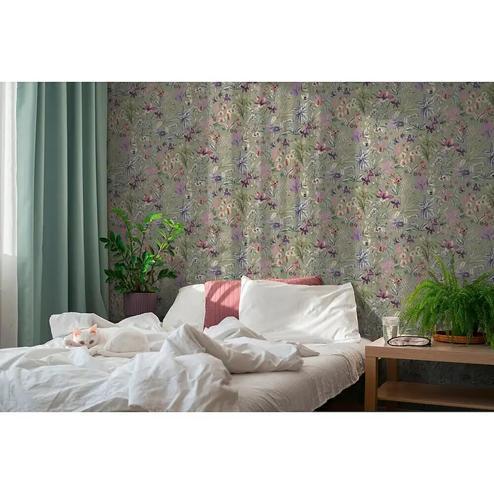 A.S. Création Pintwalls 3 Vliestapete Blüten - Grün-Lila, Floral, 10,05 x 0,53 m - Florale Tapete in Grün-, Violett- und Rosatönen schmückt eine Schlafzimmerwand hinter einem Bett mit einer weißen Katze.