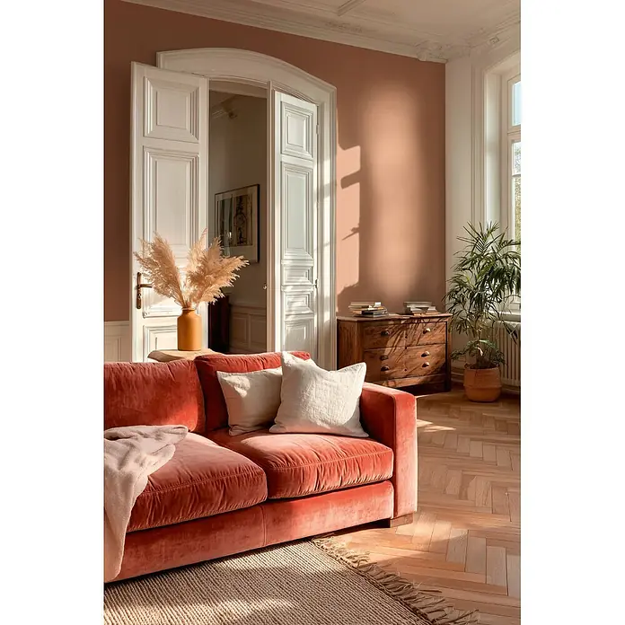 A.S. Création Metropolitan Stories Vibes & Styles Vliestapete Uni I - Rotbraun, Uni, 10,05 x 0,53 m - Ein plüschiges korallenrotes Samtsofa mit hellen Kissen und einer Decke in einem sonnigen Wohnzimmer, das sein bequemes Design hervorhebt.