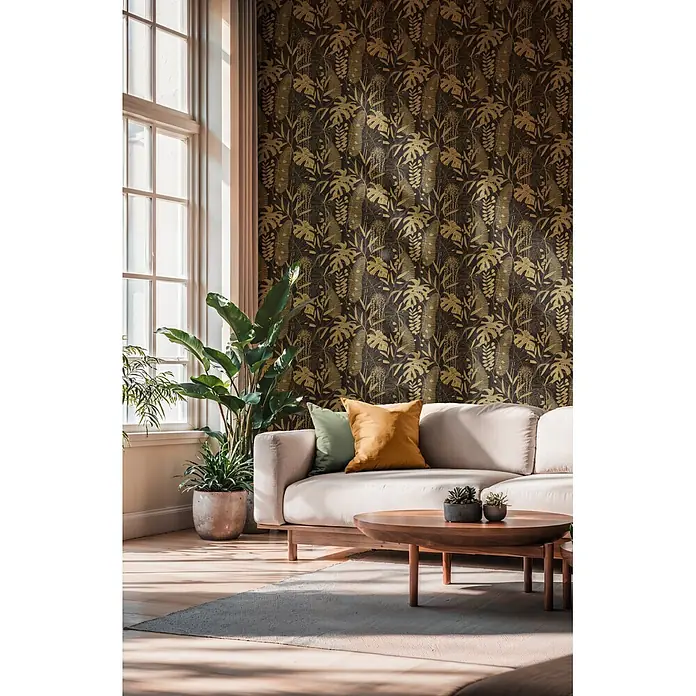 A.S. Création Cuba 2 Vliestapete Blätter - Schwarz-Gold, Floral, 10,05 x 0,53 m - Wohnzimmerwand mit dunkler Tapete mit goldenen Tropenblattmustern hinter hellem Sofa und Pflanzen.