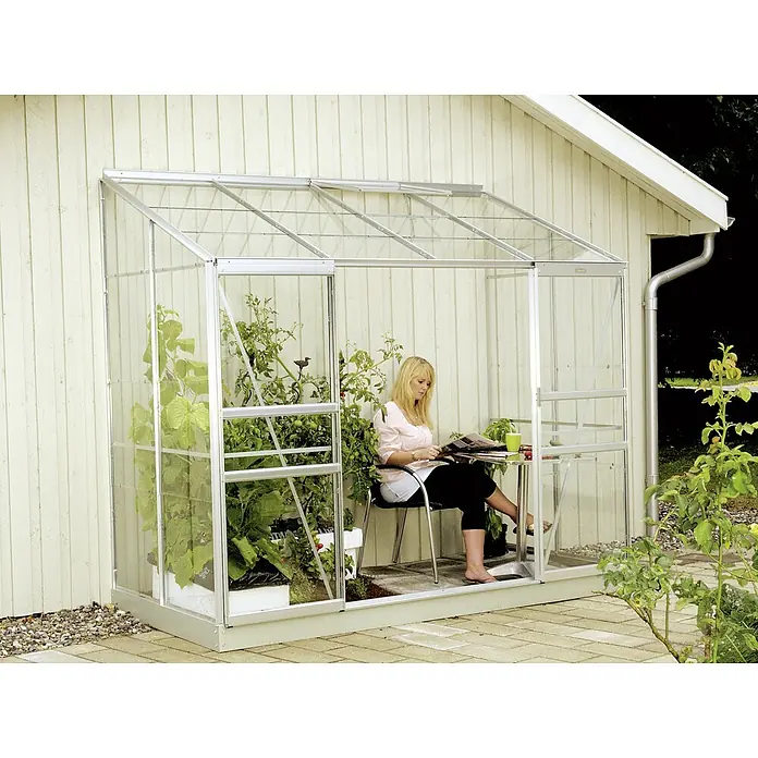 Vitavia Gewächshaus Styx 3300 - Außenmaß inkl. Dachüberstand (B x T): 255 x 132 cm, 3 mm, Aluminium - Ein Anlehngewächshaus aus Aluminium und Glas ist an eine Hauswand angebaut, eine Frau liest darin eine Zeitschrift.
