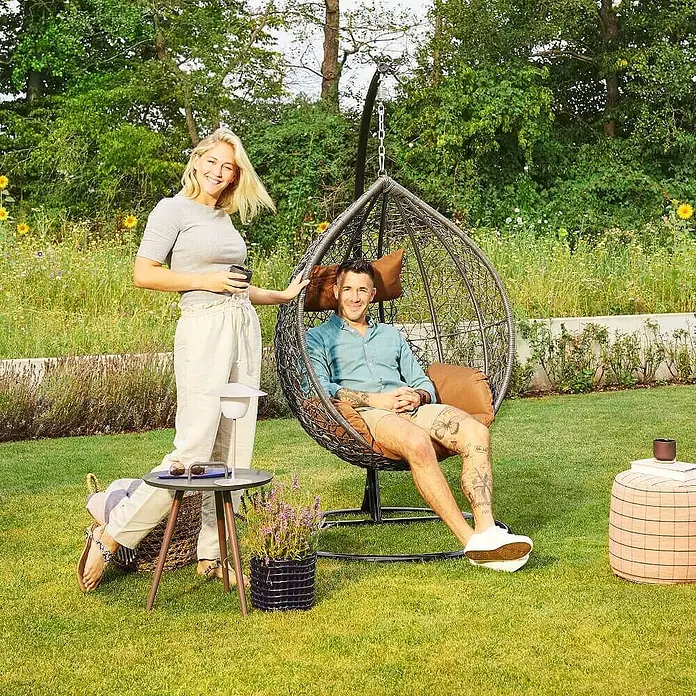 Home Deluxe Hängesessel CIELO - L x B x H: 104 x 104 x 200 cm, Grau - Ein Mann entspannt in einem dunklen Hängesessel mit braunen Kissen in einem sonnigen Garten.