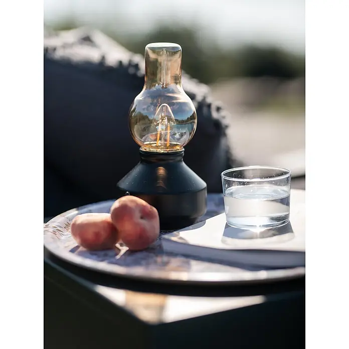 PR Home Akku-Tischleuchte schwarz Outdoor - Warmweiß, 1 W - Vintage-Tischlampe mit schwarzem Fuß und bernsteinfarbener Edison-Glühbirne auf einem Gartentisch mit Pfirsichen und Wasser.