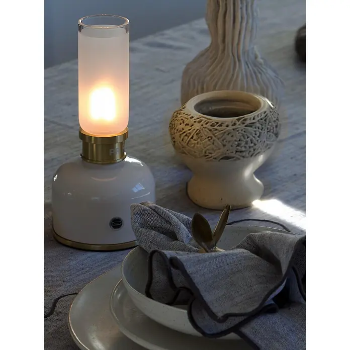 PR Home Akku-Tischleuchte mit flackerndem Licht - Warmweiß, 25 W - Weiße und messingfarbene tragbare Laterne mit mattiertem Glasschirm, beleuchtet einen Esstisch.