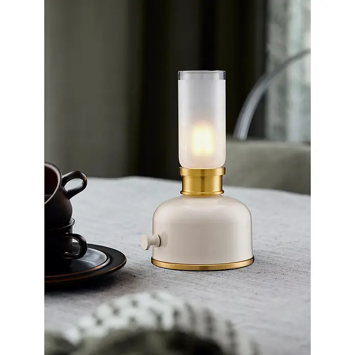 PR Home Akku-Tischleuchte Frida - Warmweiß, 0,33 W - Eine beleuchtete tragbare Lampe in Creme und Messing mit mattiertem Glasschirm steht auf einem Tisch neben gestapelten dunklen Tassen.