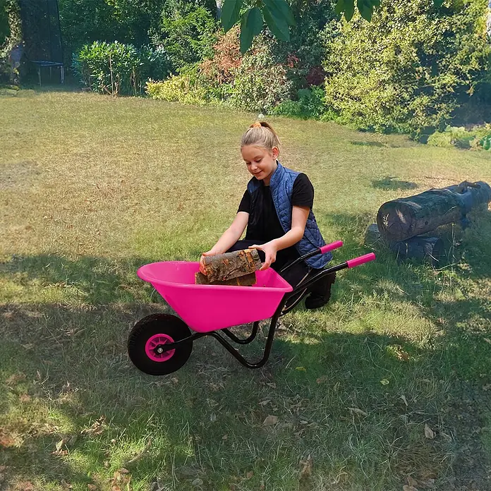 Pro-Bau-Tec Bollerwagen Kinderschubkarre Pink mit PP-Mulde 20L mit PU-Rad - L x B x H: 1,05 cm x 0,34 cm x 0,39 m - Ein Mädchen legt Holzscheite in eine leuchtend pinke Schubkarre im Garten.