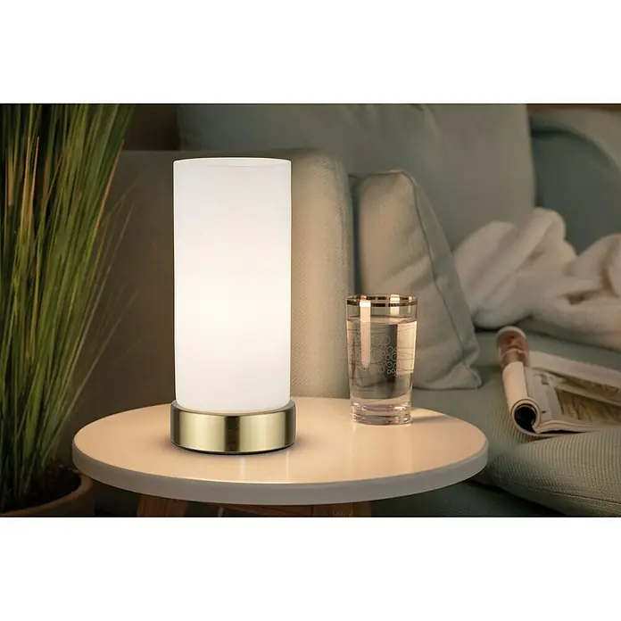 Paulmann Lámpara de sobremesa Pinja - 20 W, L x An x Al: 11 cm x 11 mm x 24,4 cm, Latón, Blanco, E14 - Primer plano de una moderna lámpara de noche con un vaso y una revista en una pequeña mesa junto a un sofá.