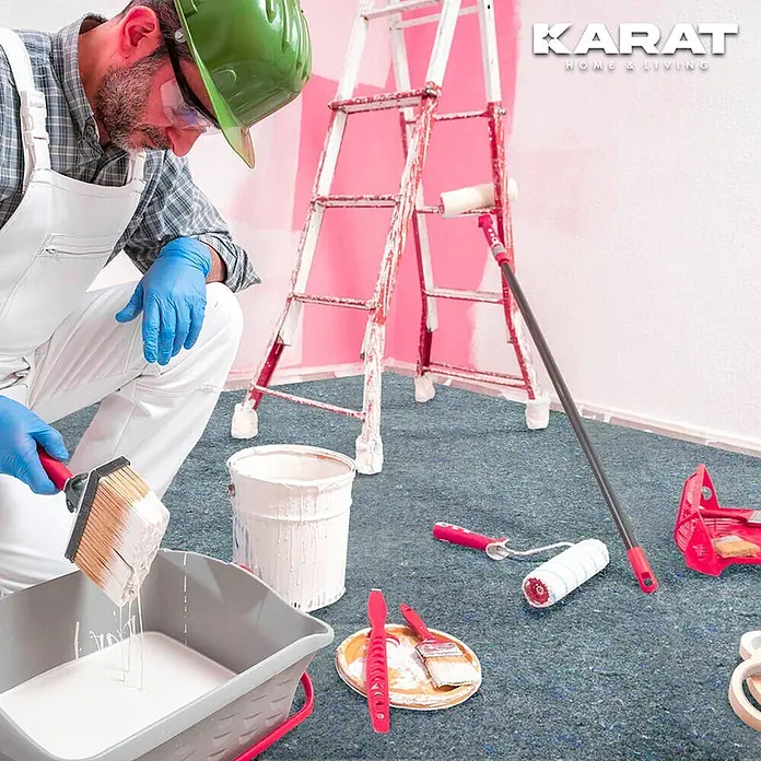 Floordirekt Abdeckvlies 180g/m² - Hochwertiger Schutzvlies für Malerarbeiten - 50 x 1 m - Maler in weißem Overall und blauen Handschuhen taucht Pinsel in weiße Farbe. Werkzeuge, Leiter und Bodenschutz sind sichtbar.
