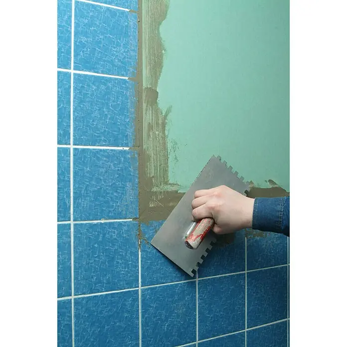 Knauf Gipskartonplatte GKBI - 1.250 x 900 x 6,5 mm, Imprägniert - Nahaufnahme einer Person, die mit einer gezahnten Kelle Fugenmasse auf blaue Fliesen aufträgt, um eine glatte und haltbare Oberfläche zu schaffen.