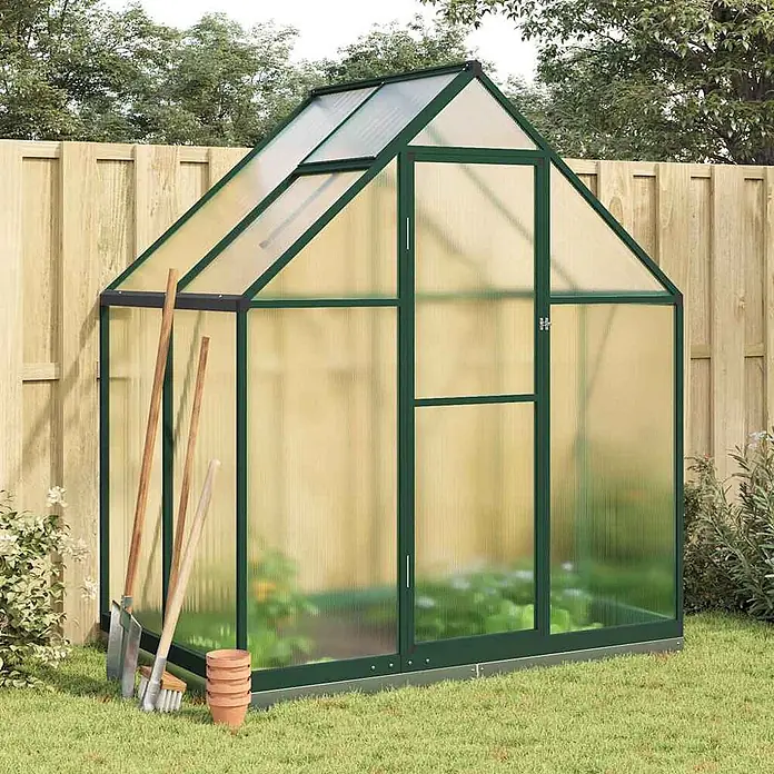 Furnicato Gewächshaus - Außenmaß inkl. Dachüberstand (B x T): 114 x 169 cm, Material Seitenwand: Aluminium, 0,4 mm, grün - Grün gerahmtes Gewächshaus mit durchscheinenden Paneelen, in einem Garten mit Pflanzen und Gartengeräten.