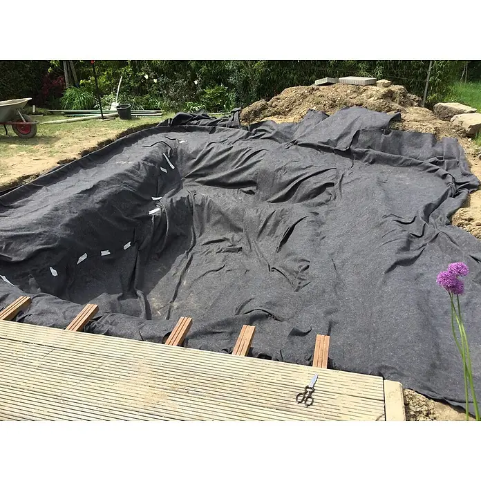 Aquagart Bodenschutzvlies 24m² 500g/m² Teichvlies Teich Schutzvlies -  - Dunkelgraues Teichvlies wird in einem Gartenteich verlegt, neben einer Holzterrasse.