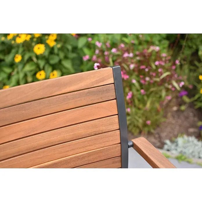 Merxx Gartenstuhl-Set Santorin - 2 Stk., Aluminium/Akazie, Stapelbar, graphit/natur - Nahaufnahme einer Gartenbank aus Holzlatten und dunklem Metallrahmen, umgeben von bunten, unscharfen Blumen.