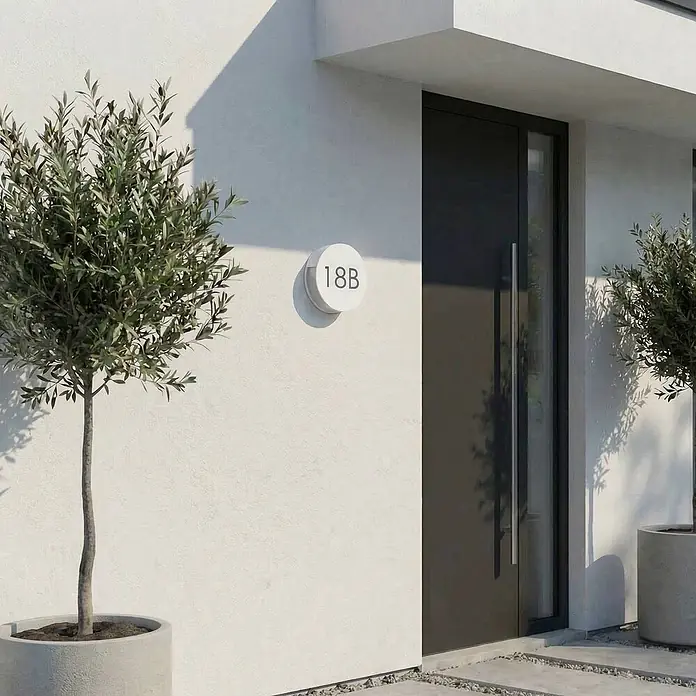 Lucande Außenwandleuchte Advero - alu / grau / zink, alu / grau / zink, IP54 - Runde weiße Hausnummer '18B' an moderner weißer Wand neben dunkler Tür, mit Olivenbäumen in Töpfen.