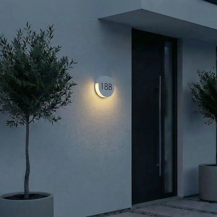 Lucande Außenwandleuchte Advero - alu / grau / zink, alu / grau / zink, IP54 - Runde weiße Außenwandleuchte, die die Hausnummer '18B' an einer modernen Fassade beleuchtet.