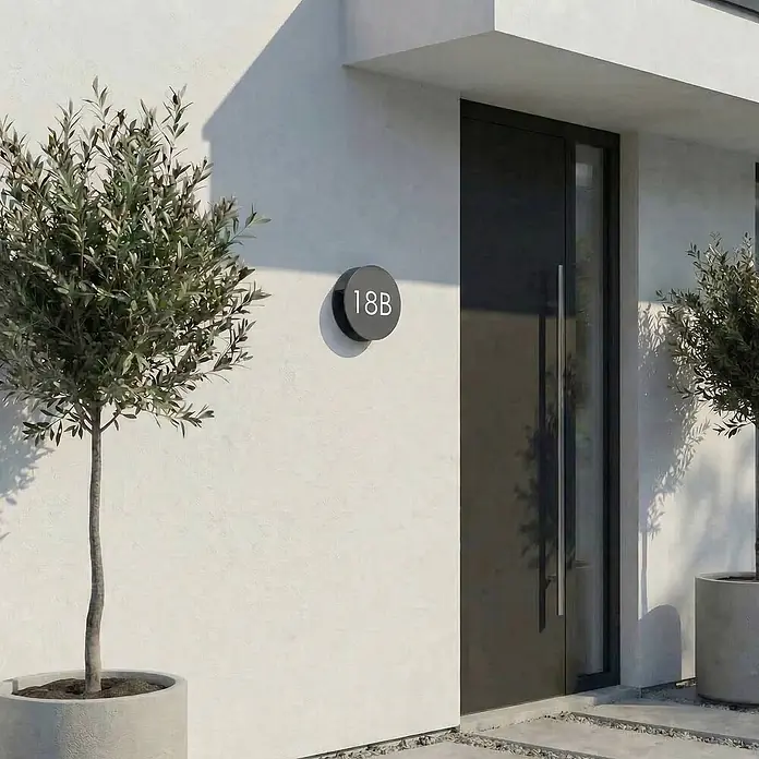 Lucande Außenwandleuchte Advero - 6 x 23 x 23 cm, schwarz, schwarz, IP54 - Rundes dunkelgraues Hausnummernschild 18B mit weißen Ziffern, an einer hellen Wand neben einem modernen Eingang mit Olivenbaum montiert.
