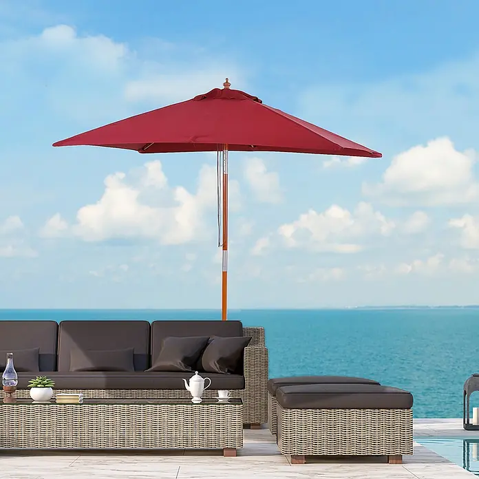 Furnicato Balkonschirm - Weinrot - Outdoor-Lounge-Set aus Rattan mit dunklen Kissen und rotem Sonnenschirm am Wasser.