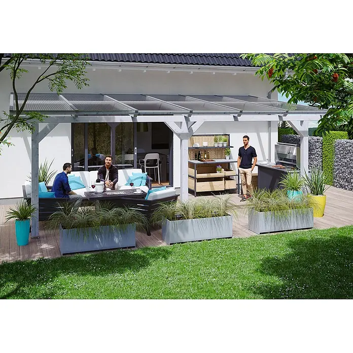 SKAN HOLZ Terrassenüberdachung Andria - B x T: 648 x 350 cm, Weiß, Polycarbonat, Transparent - Graue rechteckige Pflanzkübel mit hohem Ziergras, die eine Holzterrasse von einem grünen Rasen abgrenzen.