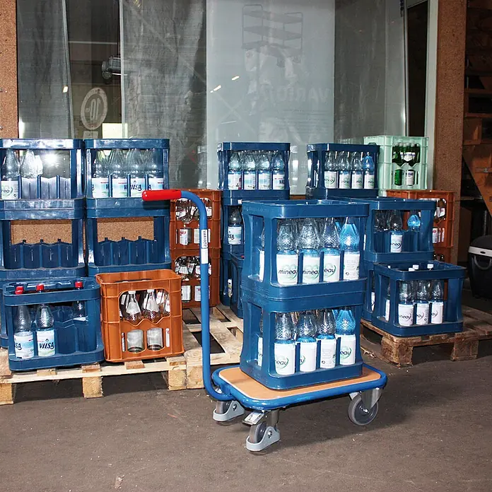 Variofit Transportroller Griffroller mit Boden in Buchendekor 605x500mm RAL 5010 Enzianblau -  - Blauer Handwagen mit Holzplattform und rotem Griff, transportiert gestapelte blaue Getränkekisten mit Flaschen in einem Lager.