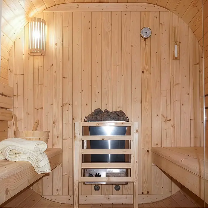 Home Deluxe Fasssauna CUBE L - Elektroofen, Holz, L x B x H: 180 x 192,8 x 194 cm - Innenansicht einer hellen Holzsauna mit Bänken, Heizgerät mit Steinen und Wandlampe, bereit zur Nutzung.