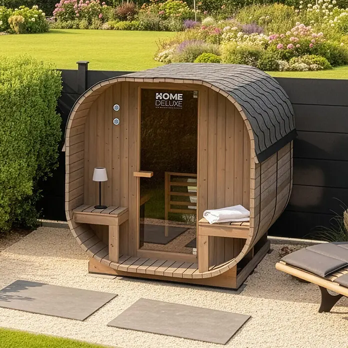 Home Deluxe Fasssauna CUBE DELUXE L - Elektroofen, Holz, L x B x H: 220 x 192,8 x 194 cm - Hölzerne Fasssauna mit dunklem Schindeldach in einem ruhigen Garten mit Kies und Pflastersteinen.
