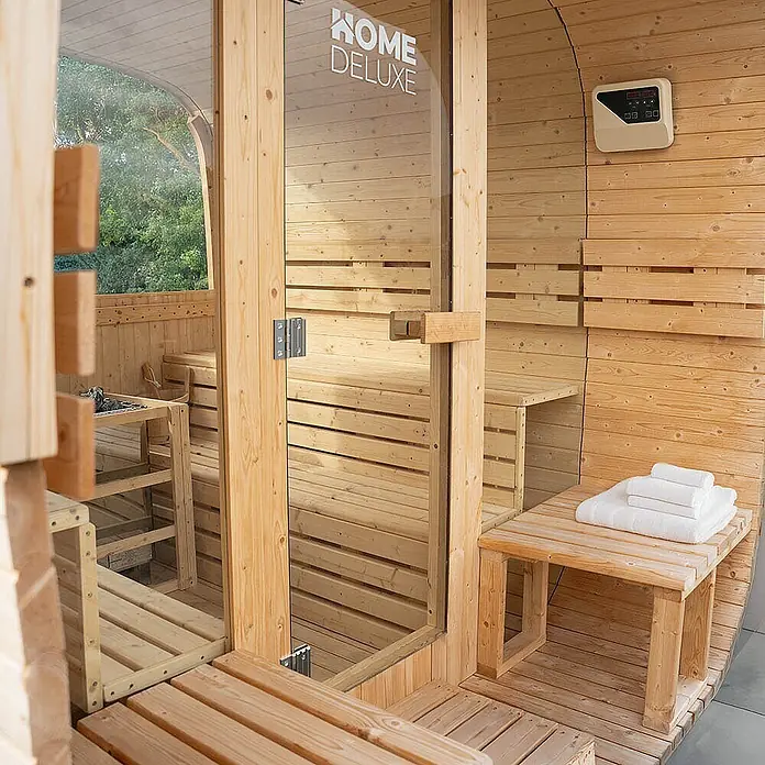 Home Deluxe Fasssauna HYGGE DELUXE Größe: 230 x 220 cm - Elektroofen, Holz, L x B x H: 260 x 230 x 220 cm - Home Deluxe Holzfasssauna mit Glastür, Innenbänken und einer Außenbank mit weißen Handtüchern.