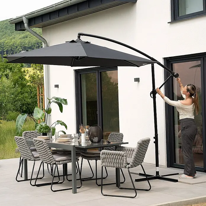 Home Deluxe Ampelschirm SOLANA - Anthrazit - Graues Outdoor-Essgruppe mit geflochtenen Stühlen und großem Ampelschirm auf einer modernen Terrasse, von einer Frau eingestellt.