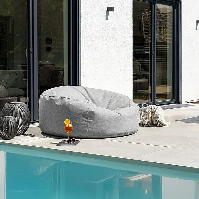 Home Deluxe Gartenhocker CLOUD Pouf - Hellgrau, B x T x H: 140 x 140 x 40 cm - Großer grauer Outdoor-Sitzsack auf einer Terrasse am Pool, mit Getränk und Sonnenbrille.