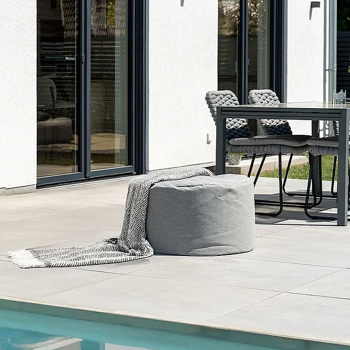 Home Deluxe Gartenhocker CLOUD Pouf - Hellgrau, B x T x H: 70 x 70 x 40 cm - Grauer runder Outdoor-Pouf mit gemusterter Decke, auf einer Terrasse neben einem Pool.