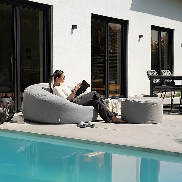 Home Deluxe Gartenhocker CLOUD Pouf Hocker-Set - Hellgrau, B x T x H: 140 x 140 x 40 cm - Eine Frau entspannt auf einem großen grauen Sitzsack neben einem Pool, mit passendem Hocker, auf der Terrasse.