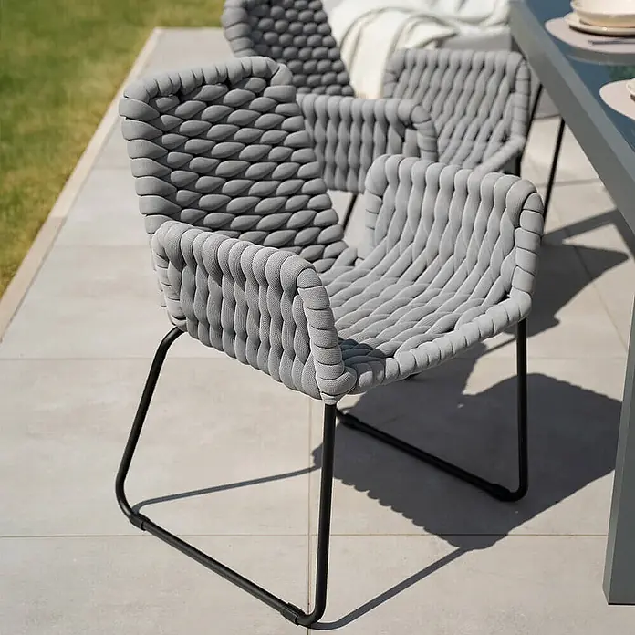 Home Deluxe Gartenstuhl mit Armlehnen BALERO - 7er Set - Pulverbeschichtet, Hellgrau, B x T x H: 63 x 62 x 85 cm - Graue Outdoor-Stühle aus Geflecht mit schwarzen Metallbeinen auf einer Terrasse.