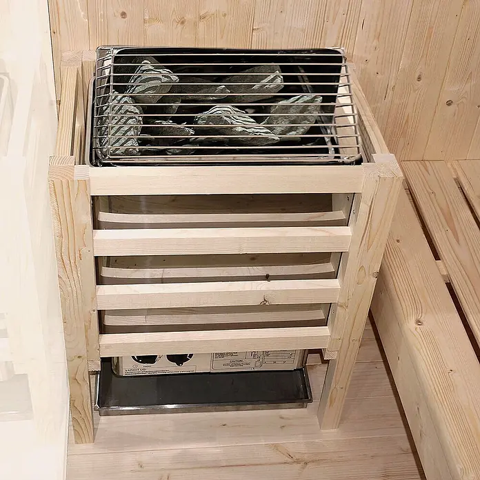 Home Deluxe Elementsauna SHADOW XL - Elektroofen, Steuerung Ofen: Integriert - Saunaofen mit Steinen, Metallgittern und einem Schutzgitter aus hellem Holz, in einer Sauna.