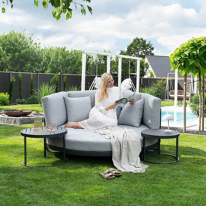 Home Deluxe Gartenliege FLORIDA - Farbe: Grau, Ausführung: nur Daybed - B x T: 170 x 20 cm, Grau, Aluminium - Rundes graues Outdoor-Daybed mit lesender Frau, flankiert von zwei kleinen dunklen Beistelltischen.