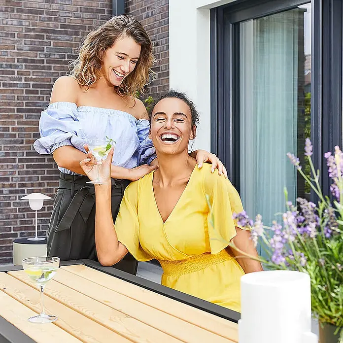 Home Deluxe Gartentisch-Set KOBON - Gesamtumfang: 3 -tlg. - Zwei Frauen lachen und genießen Getränke an einem Terrassentisch mit Holzlatten und dunklem Rahmen.