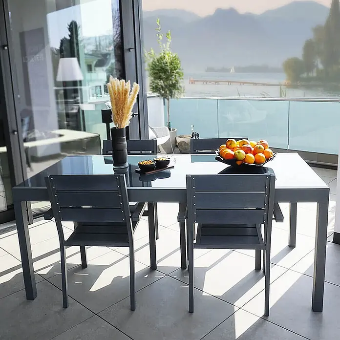 Home Deluxe Gartentisch SOL - L x B x H: 70 x 180 x 240 cm, Anthrazit - Dunkelgraue Outdoor-Essgruppe mit Glastisch und Stühlen auf einem sonnigen Balkon mit Seeblick.