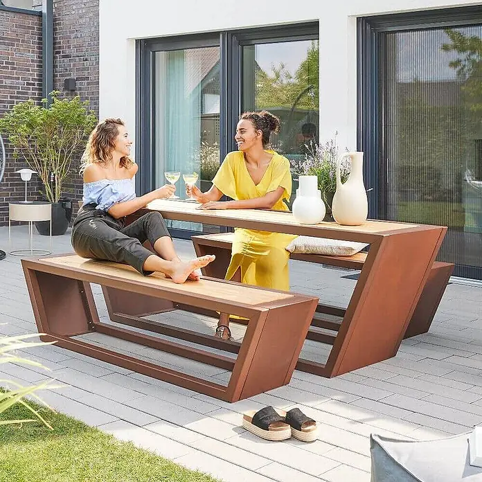 Home Deluxe Gartentisch-Set KOBON - 2x Bank + 1x Tisch - Gesamtumfang: 3 -tlg. - Outdoor-Essgruppe mit braunem Metallgestell und Holzplatten, von zwei Frauen auf einer Terrasse genutzt.