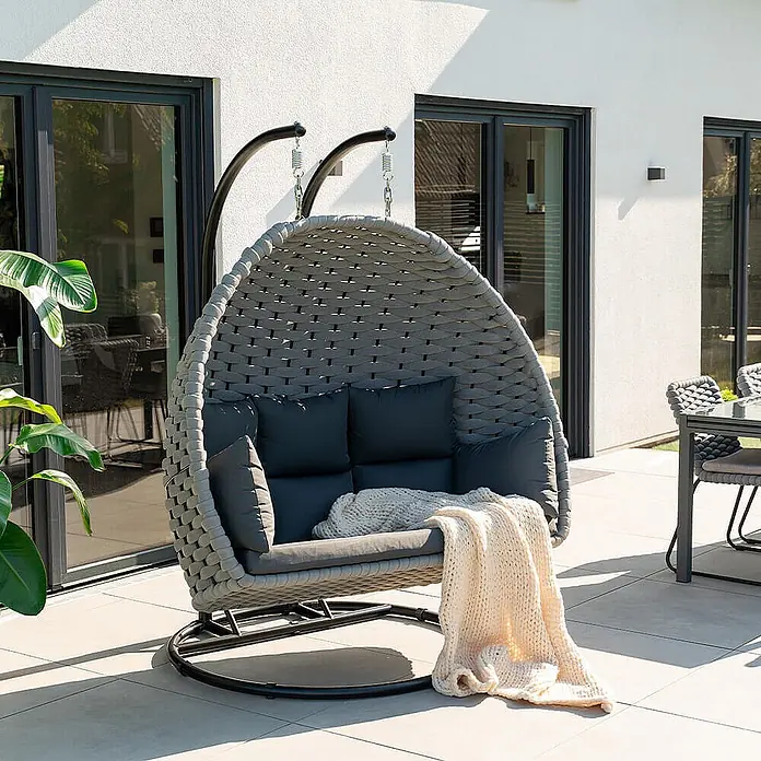 Home Deluxe Hängesessel CADEIRA 2-Sitzer - Stahl, Hellgrau - Grauer, geflochtener Hängesessel für zwei mit dunklen Kissen und einer cremefarbenen Decke auf der Terrasse.
