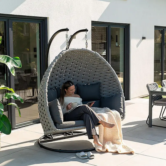 Home Deluxe Hängesessel CADEIRA 2-Sitzer - Stahl, Hellgrau - Grauer geflochtener Ei-Schaukelstuhl mit dunklen Kissen, eine Frau entspannt auf der Terrasse.