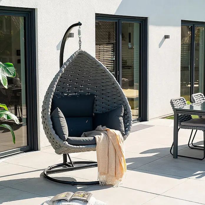 Home Deluxe Hängesessel CADEIRA 1-Sitzer - Stahl, Hellgrau - Grauer geflochtener Hängesessel mit dunklen Kissen und orangefarbener Decke auf einer sonnigen Terrasse für Entspannung.