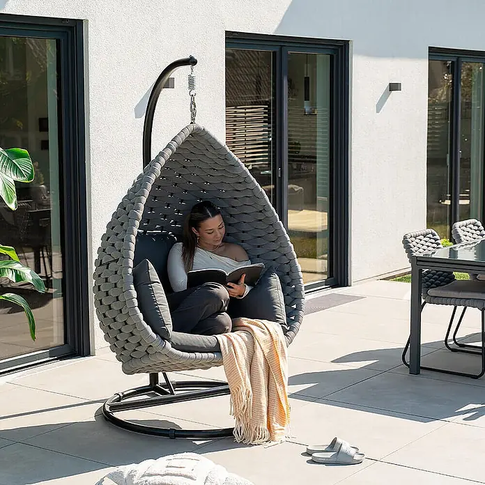 Home Deluxe Hängesessel CADEIRA 1-Sitzer - Stahl, Hellgrau - Grauer geflochtener Eier-Hängesessel mit dunklen Kissen auf einer Terrasse, von einer lesenden Frau genutzt.