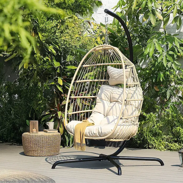 Home Deluxe Hängesessel LOMA 1-Sitzer - Kunststoff, Beige - Beiger Rattan-Hängesessel mit cremefarbenen Kissen und schwarzem Metallgestell auf einer Holzterrasse, umgeben von Grünpflanzen.