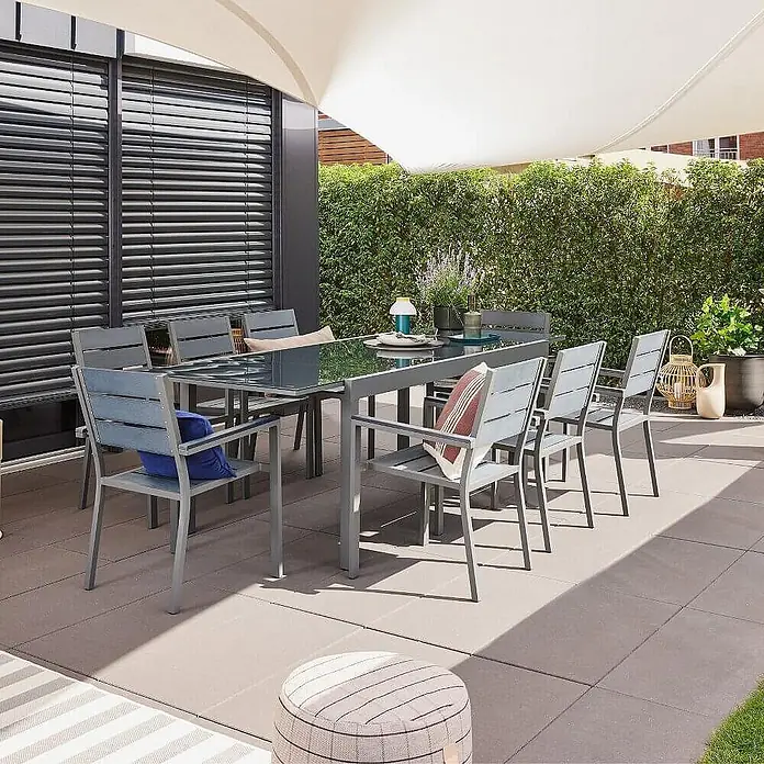 Home Deluxe Gartentisch SOL - L x B x H: 70 x 180 x 240 cm, Anthrazit - Modernes graues Outdoor-Essset mit Glastisch und Stühlen mit blauen Latten auf einer Terrasse unter einem Sonnensegel.