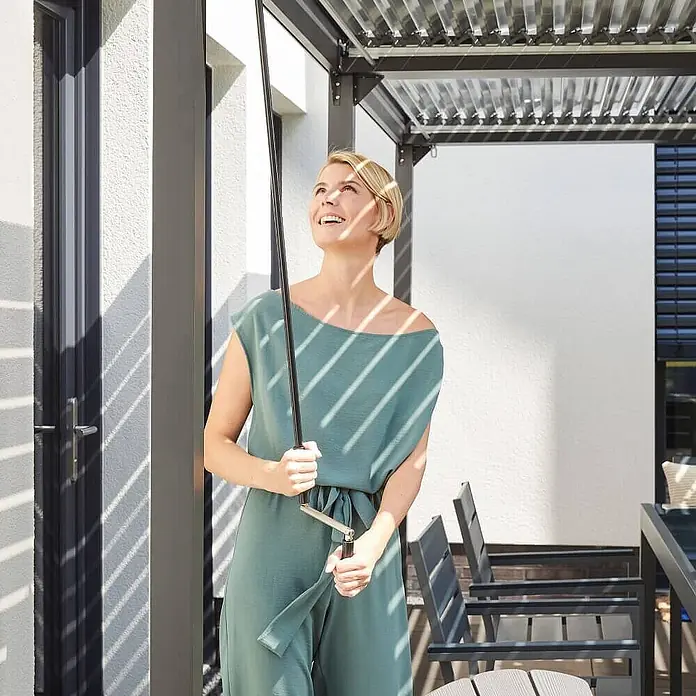 Home Deluxe Pergola CANNES - B x T: 400 x 303 cm, Farbe Gestell: Anthrazit, Aluminium - Frau bedient mit dunkler Kurbelstange eine Pergola-Lamellenüberdachung.