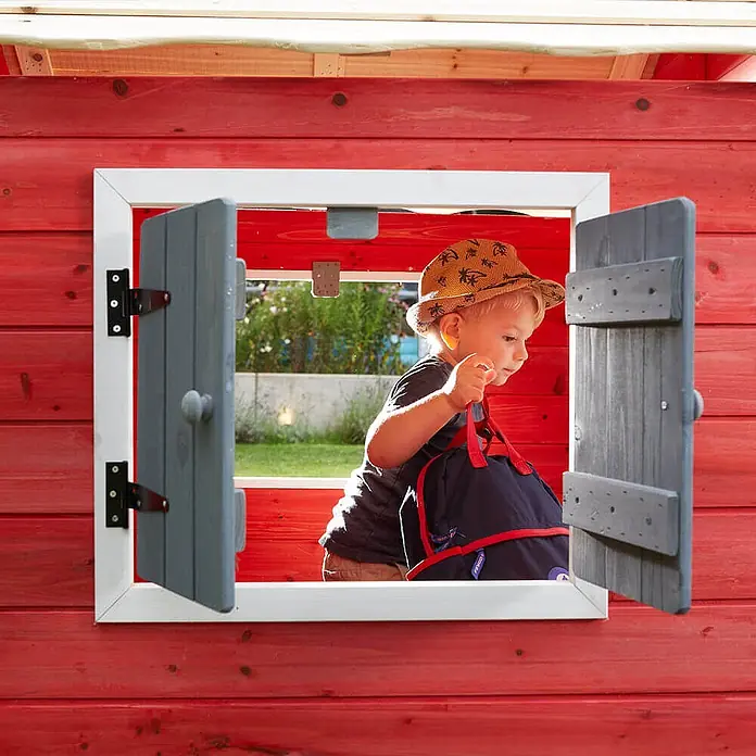 Home Deluxe Spielhaus DAS KLEINE SCHLOSS - Holz, Rot - Kind spielt in einem roten Holzspielhaus mit grauen Fensterläden und trägt einen dunkelblauen Rucksack.