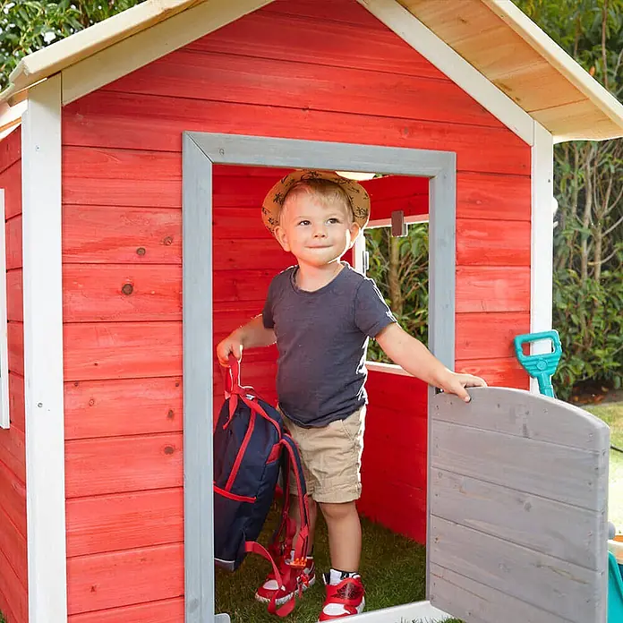 Home Deluxe Spielhaus DAS KLEINE SCHLOSS - Holz, Rot - Kleinkind steht mit Rucksack in der Tür eines roten Holzspielhauses mit grauen Zierleisten.