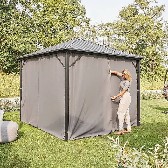 Home Deluxe Standard-Pavillon Seitenwand Vorhang AZUR - B x T: 270 x 270 cm, Farbe Gestell: Schwarz, Kunststoff - Eine Frau schließt die graue Seitenwand eines Gartenpavillons mit dunkelgrauem Dach.