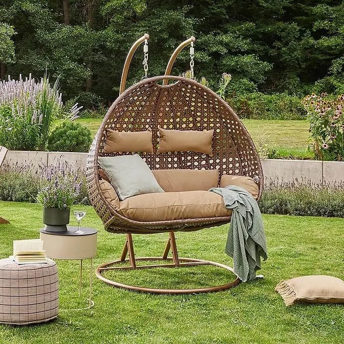 Home Deluxe Hängesessel TWIN - Kunststoff, Braun - Brauner Rattan-Doppel-Hängesessel mit beigen Kissen und Decke in einem üppigen Garten.