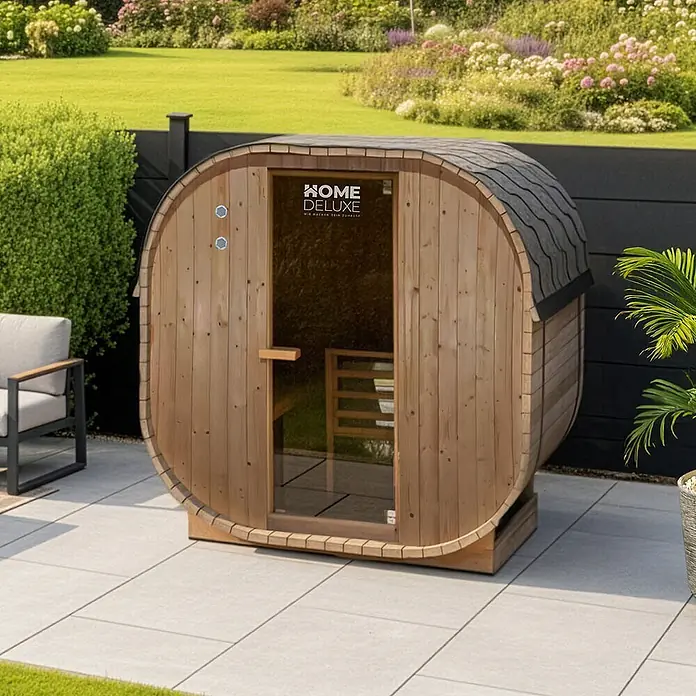 Home Deluxe Fasssauna CUBE M - Elektroofen, Holz - Hölzerne Fasssauna mit Glastür auf einer hellgrauen Terrasse in einem Garten.