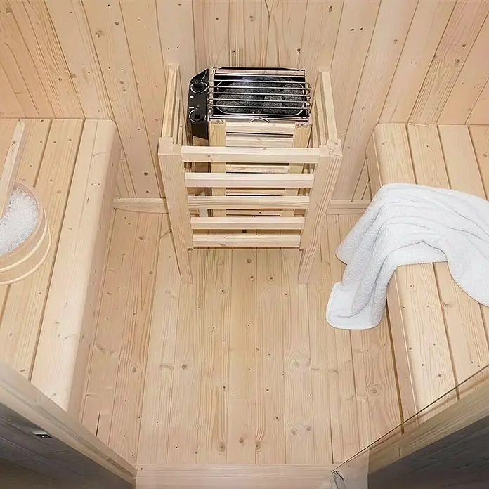 Home Deluxe Fasssauna CUBE M - Elektroofen, Holz - Heller Saunainnenraum aus Holz mit Elektroofen, zwei Bänken, einem Eimer und einem weißen Handtuch.