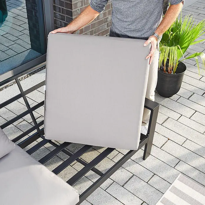 Home Deluxe Garten-Loungesessel RIO S -  - Person platziert ein großes, hellgraues quadratisches Kissen auf ein schwarzes Metall-Gartensofa.